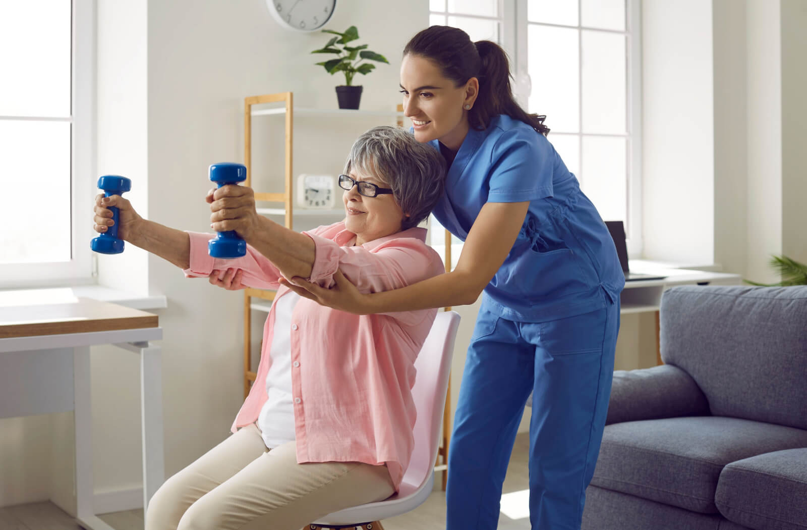 A caregiver smiling while helping a resident lift two small weights during a seated physical therapy exercise.