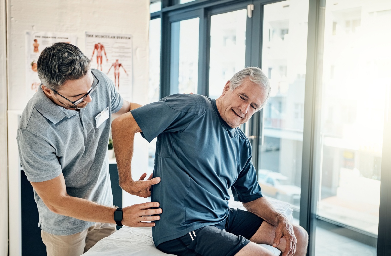 An older adult sitting on a physiotherapy table gesturing to their back while a therapist examines them.