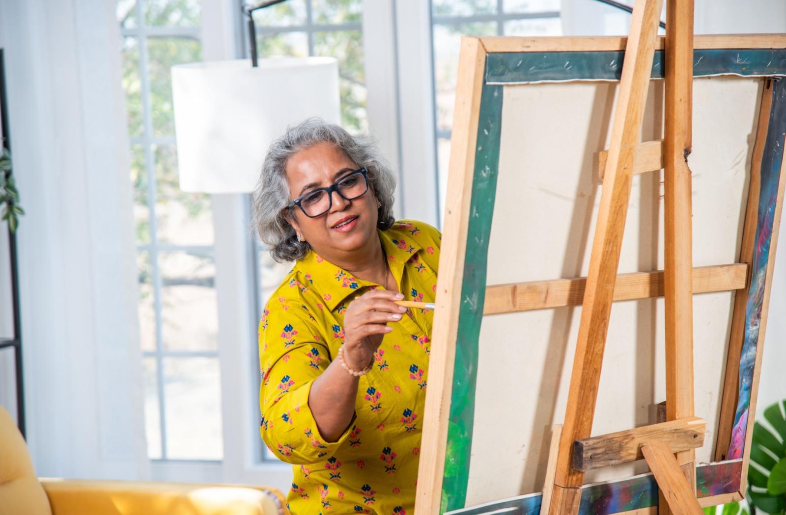 A senior in a yellow shirt uses a paintbrush to paint on a canvas set up on an easel in their well-lit living room