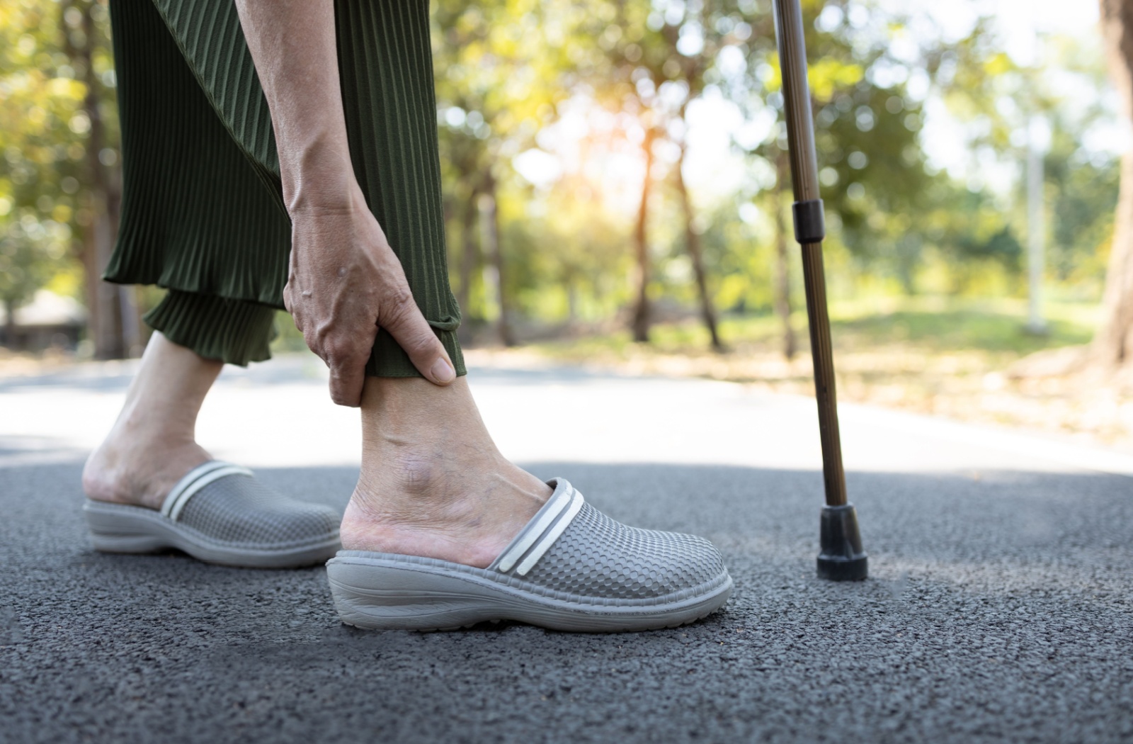 An older adult with a cane rubs their ankle while walking through a park in a pair of comfortable gray slip-on shoes