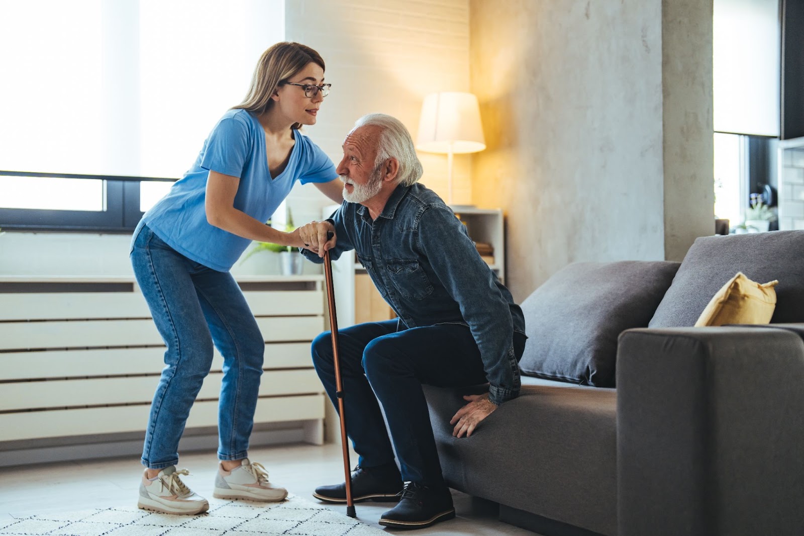 A caregiver gives a senior assistance getting to their feet from a couch.