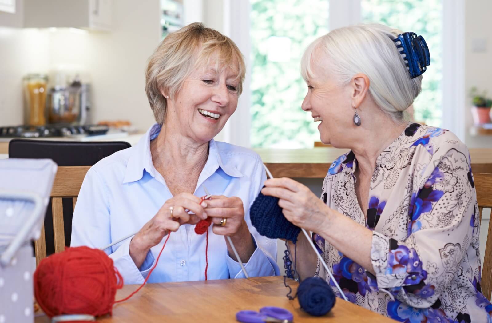 Two senior women sitting at a table knitting and smiling