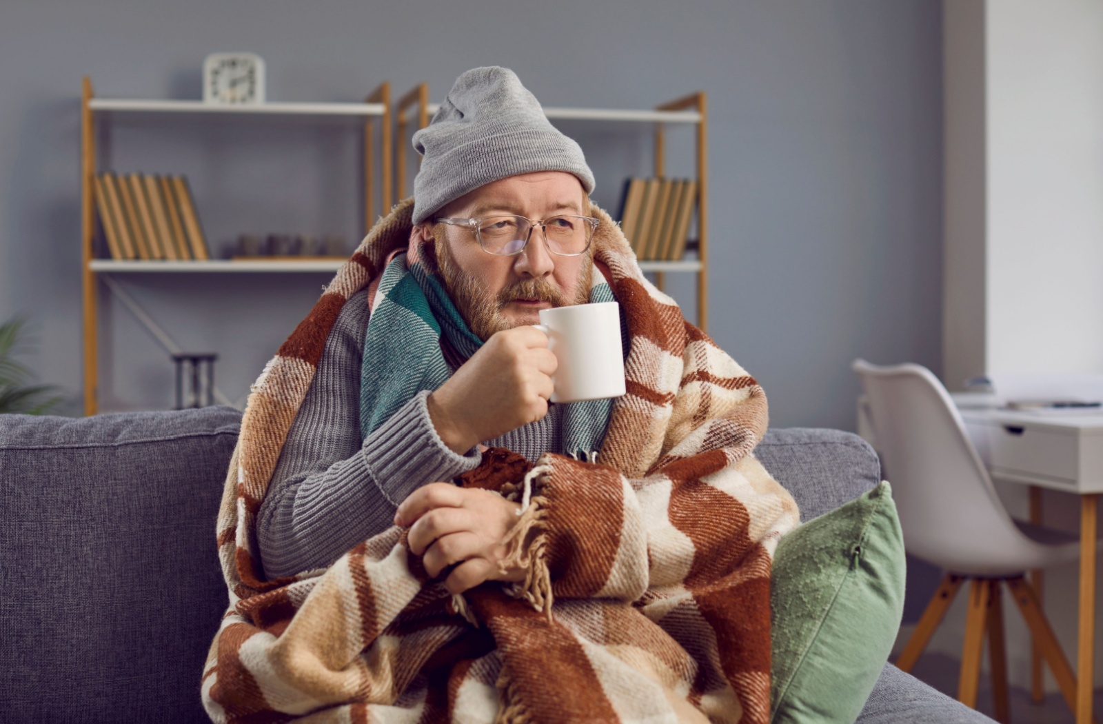 An older adult sitting on a couch wearing a scarf, hat, and blanket and holding a mug in their right hand.