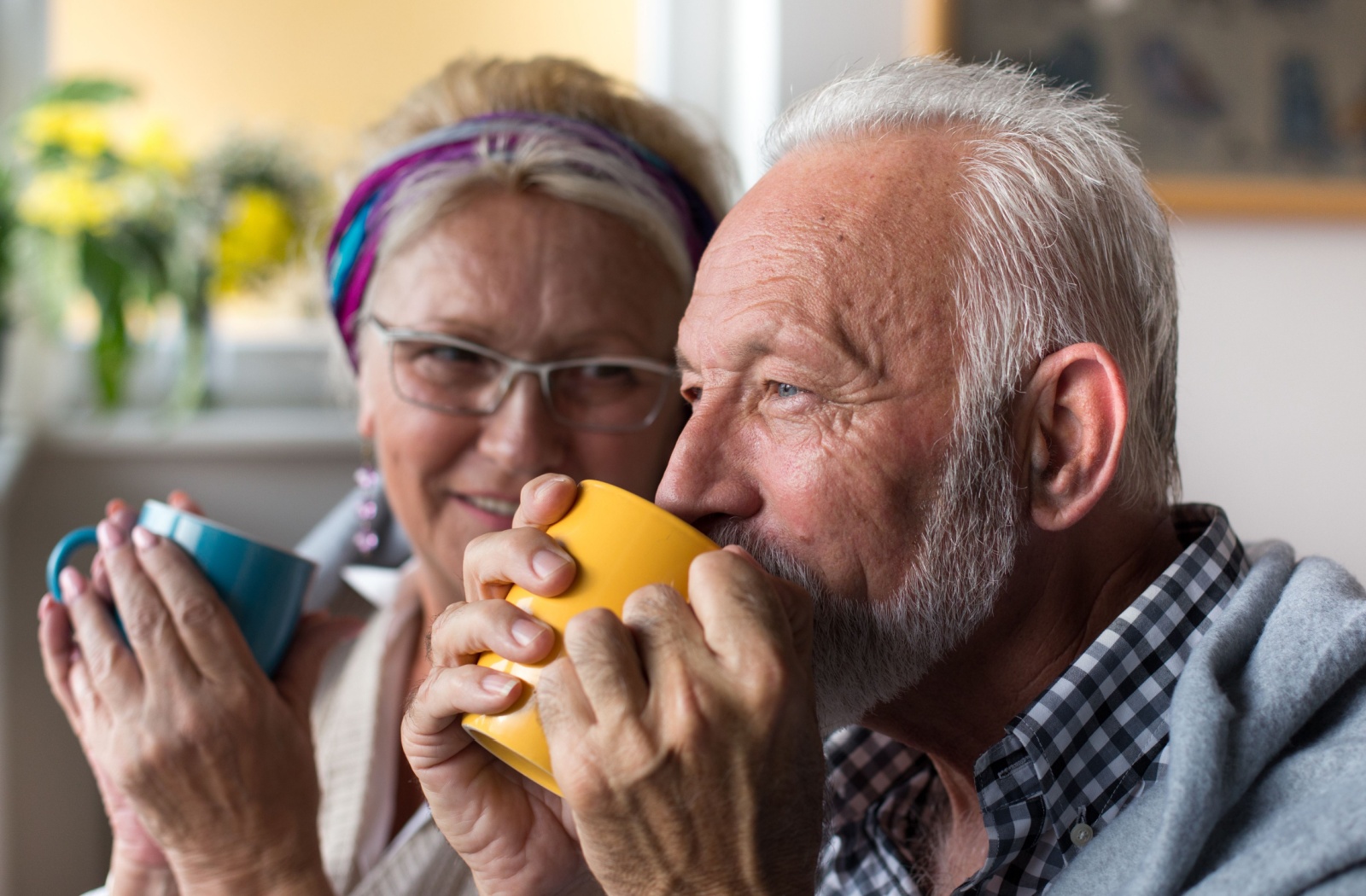 A close-up image of an older couple wrapped in a blanket and holding mugs in their hands.
