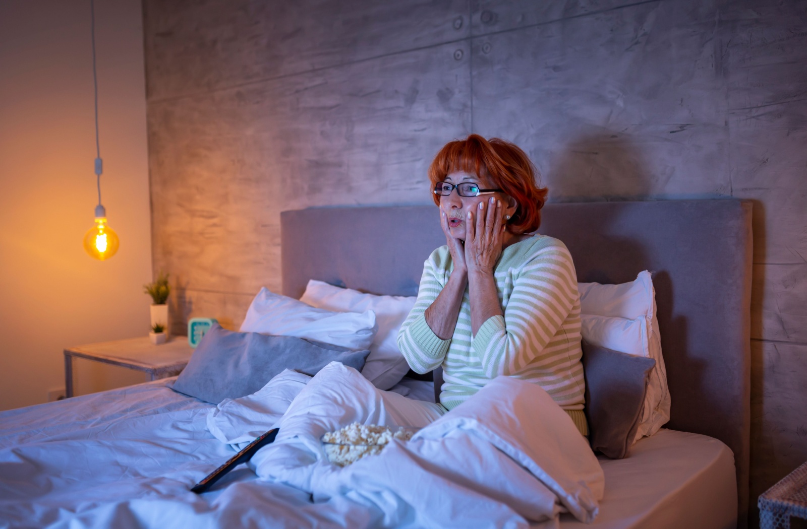 An older adult touches their face in surprise while sitting in a dim modern bedroom at night, watching a movie about Alzheimer's disease with popcorn in bed