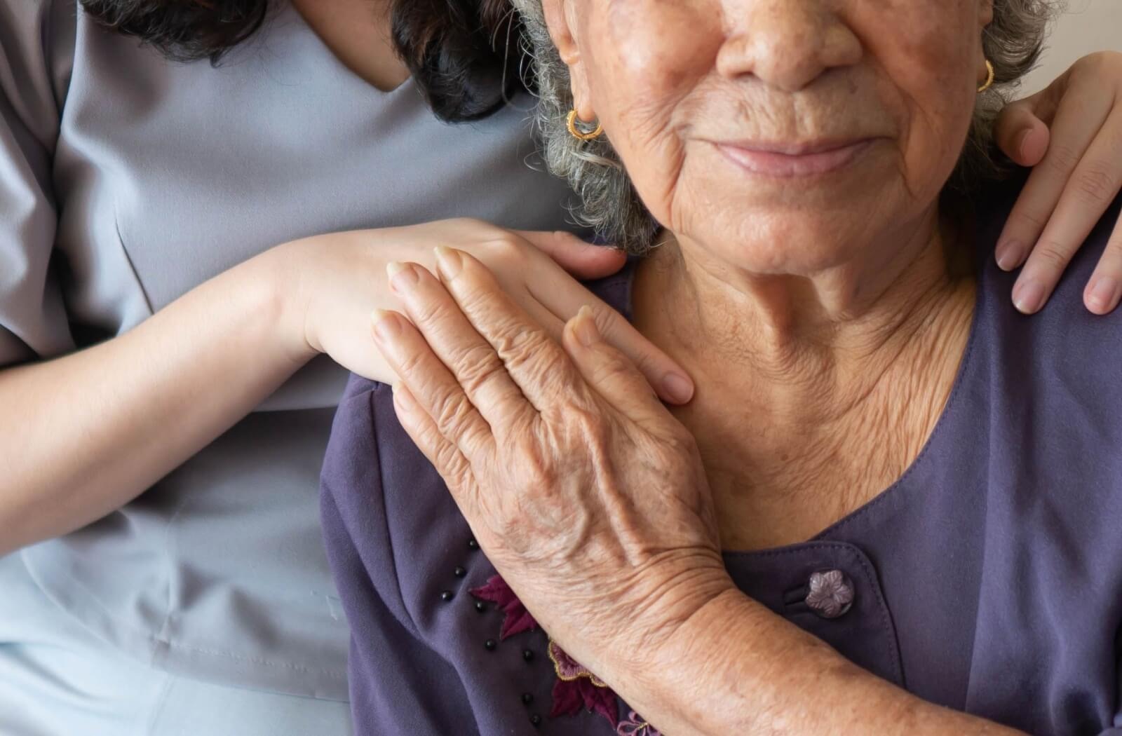 A resident in assisted living reaches up to touch the hand of a caregiver standing behind them